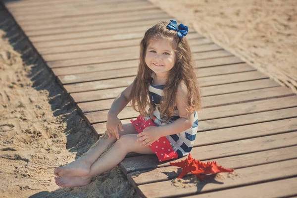 Aranyos kis lány élvezi a nyári időszakban a sea side beach boldog játszik a vörös csillag és apró játék horgony a homok nobby ruhát barna hajat visel — Stock Fotó