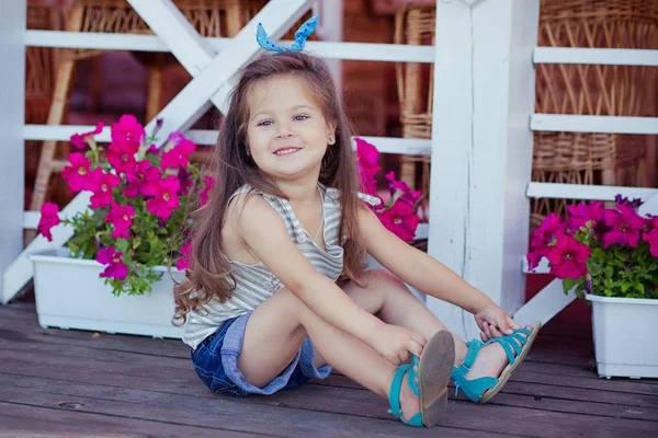 Élégant beau bébé fille mignonne avec des cheveux bruns posant sur un jardin en bois plein de fleurs portant de minuscules chemises en jeans et des sandales bleues et une taille skivy aérée — Photo