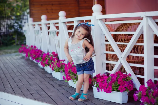 Stilvolle schöne süße Baby-Mädchen mit brünetten Haaren posiert auf hölzernen Garten voller Blumen tragen winzige Jeans-Shirts und luftige skivy Untertaille und blaue Sandalen — Stockfoto