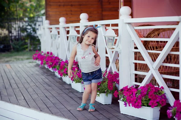 Élégant beau bébé fille mignonne avec des cheveux bruns posant sur un jardin en bois plein de fleurs portant de minuscules chemises en jeans et des sandales bleues et une taille skivy aérée — Photo