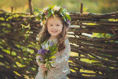 Cute young girl with brown eyes brunnette plait hair and pink cheek wearing white dress shirt and posing close to wooden house fence wagon looking to camera with wreath of flowers on head clipart