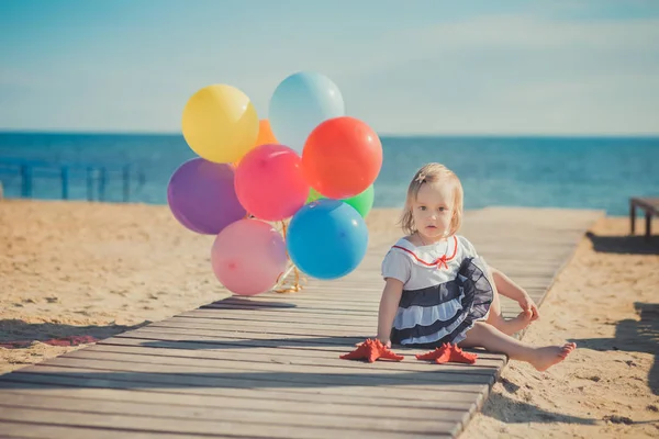 Söt blond baby flicka barn poserar njuter sommaren livstid på sandstranden havet sida på träpiren med färgglada ballonger gott bär casual klänning barfota — Stockfoto