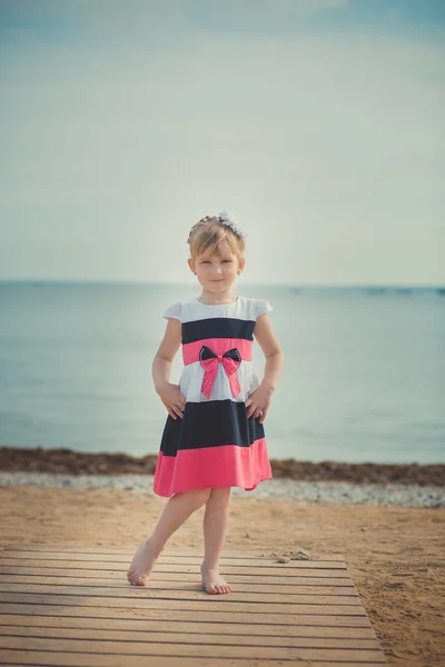 Fiatal aranyos baba lány élvező gyermekkori nyáron homokos strandon, pózol a fa mólón híd alkalmi elegáns ruha viselése — Stock Fotó
