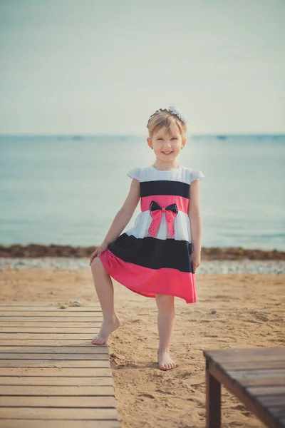 Jonge schattige babymeisje genieten van jeugd zomertijd aan zandstrand poseren op houten pier brug dragen casual stijlvolle kleding — Stockfoto