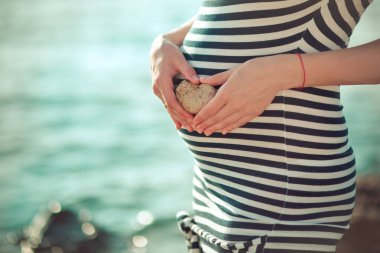 Hamile genç bayan gelecekte anne önünde poz beach Rıhtımlar üzerinde şık siyah-beyaz elbise onun karın doğmamış bebek çocuk üzerinde holding ihale eller taş kalp ile fotoğraf makinesinin odak