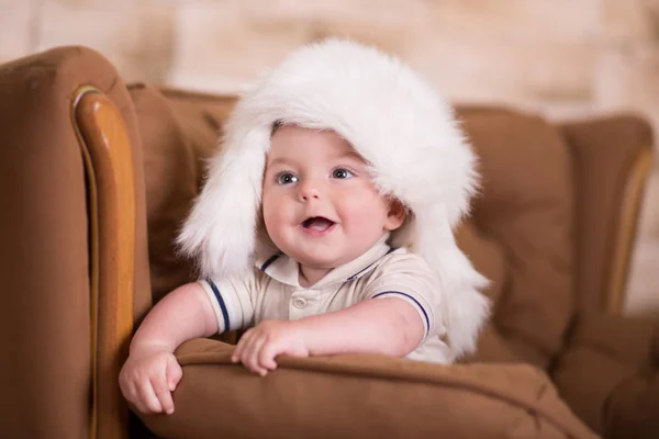Szczęśliwy nowo narodzonego dziecka chłopca z różowymi policzkami, pozowanie na hege retro styl casual brązowej kanapie divan sofa z duże białe futro zimą ciepła Czapka — Zdjęcie stockowe