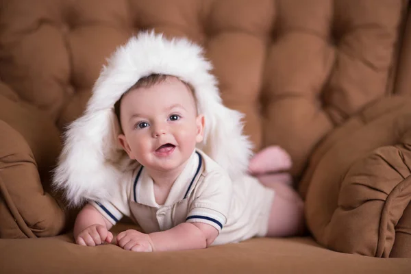 Szczęśliwy nowo narodzonego dziecka chłopca z różowymi policzkami, pozowanie na hege retro styl casual brązowej kanapie divan sofa z duże białe futro zimą ciepła Czapka — Zdjęcie stockowe