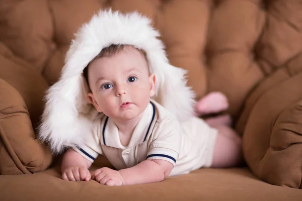 Gott nyfött barn pojke med rosa kinder poserar på hege retro casual stil brun soffa divan soffa med stora vita vintern varm pälsmössa — Stockfoto