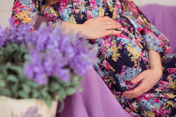 Pregnant lady woman posing sitting on sofa divan with violet balnket close to bouquet of lavender spring wild flowers wearing colourful stylish dress and holding her tummy unborn baby child — Stock Photo, Image