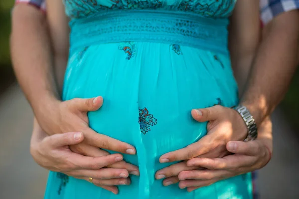Pregnant lady woman posing with her husband in front focus of camera holding by tender hands her his their tummy unborn baby child and dreaming about bright happy future wearing stylish blue dress — Stock Photo, Image