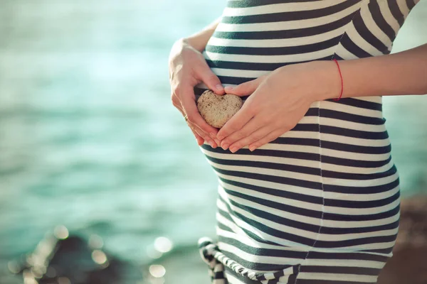 Hamile genç bayan gelecekte anne önünde poz beach Rıhtımlar üzerinde şık siyah-beyaz elbise onun karın doğmamış bebek çocuk üzerinde holding ihale eller taş kalp ile fotoğraf makinesinin odak