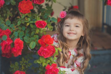 Şirin Bebek kız sarışın kıvırcık saçlar ve mutlu parlak çocuk gözleri central Park'ta çiçek büyük bush yakın kırmızı beyaz pembe Gül yaz şık kıyafetlerle poz.