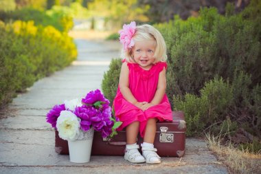 Amazing deep blue eyes baby girl child stylish dressed in colourful pink dress with shining blond hairs and white sandals posing sit for camera summer central park forest meadow with flowers and trunk clipart