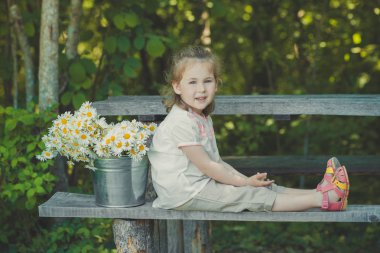 Sevimli sarışın genç kız çocuk şık beyaz gömlek ve kova papatya papatya mayweed tam poz tahta bankta oturan kısa pantolon giymiş. Adoreable sahnesi