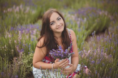 Beautifull brunette buxom nubile lady woman with deep blue eyes posing on field of spring lavender wearing cute cozy white hand made dress and holding basket with summer flowers clipart