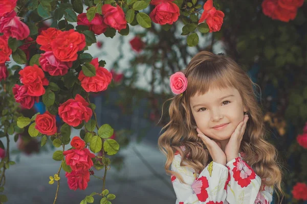 Carino bambina con i capelli biondi ricci e felici occhi luminosi bambino in posa in Central Park vicino a enorme cespuglio di fiori rosso bianco rosa rose che indossano vestiti alla moda estate — Foto Stock