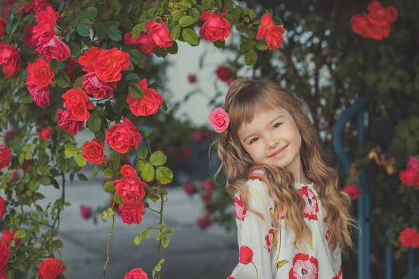 Roztomilá dívka s blond kudrnaté vlasy a očima šťastné světlé dítě pózuje v central parku blízko obrovské bush květů červené bílé růžové růže stylové oblečení letní — Stock fotografie