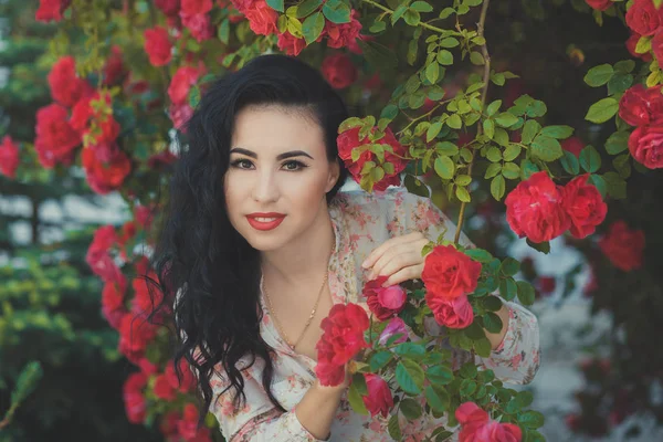 Amazing mystic lady woman with black dark curly hairs pink cheeks pout deep red lips wearing designer couturier shirt and blue short jeans shorts with brown belt posing sit for bush of flowers roses