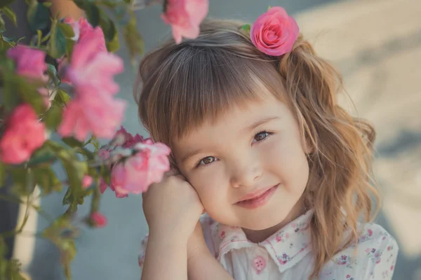 Carino bambina con i capelli biondi ricci e felici occhi luminosi bambino in posa in Central Park vicino a enorme cespuglio di fiori rosso bianco rosa rose che indossano vestiti alla moda estate — Foto Stock