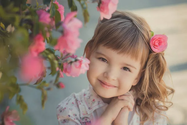 Carino bambina con i capelli biondi ricci e felici occhi luminosi bambino in posa in Central Park vicino a enorme cespuglio di fiori rosso bianco rosa rose che indossano vestiti alla moda estate — Foto Stock