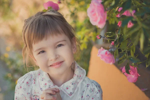 Carino bambina con i capelli biondi ricci e felici occhi luminosi bambino in posa in Central Park vicino a enorme cespuglio di fiori rosso bianco rosa rose che indossano vestiti alla moda estate — Foto Stock