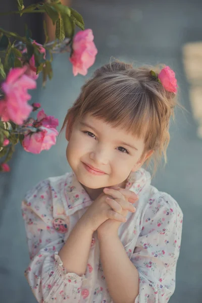 Мила дівчинка з світлим кучерявим волоссям і щасливими яскравими дитячими очима позує в центральному парку близько до величезного куща квітів червоних білих рожевих троянд в літньому стильному одязі — стокове фото