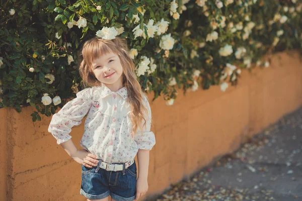 Menina bonito com cabelos cacheados loiros e felizes olhos de criança brilhante posando no parque central perto de enorme arbusto de flores rosas brancas vermelhas rosa vestindo roupas elegantes verão — Fotografia de Stock