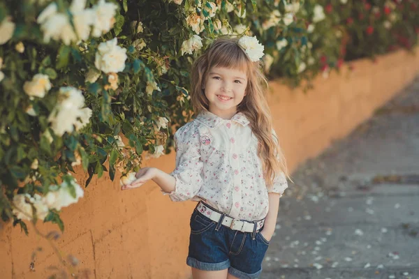 Cute baby girl with blond curly hairs and happy bright child eyes posing in central park close to huge bush of flowers red white pink roses wearing summer stylish clothes — Stock Photo, Image