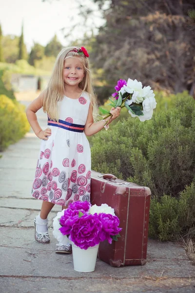 令人惊叹的深蓝色的眼睛婴儿女孩时尚穿着五颜六色的粉红色的礼服与闪亮的金发和白色凉鞋摆在相机夏天中央公园森林草地与花卉和树干 — 图库照片