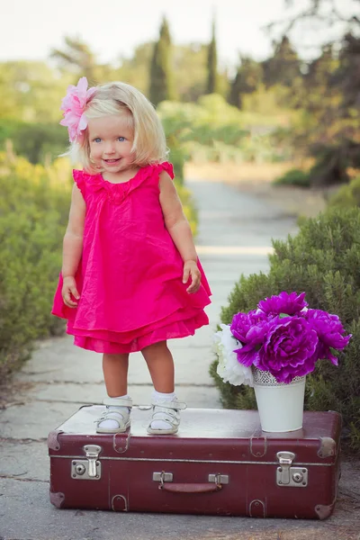 令人惊叹的深蓝色的眼睛婴儿女孩时尚穿着五颜六色的粉红色的礼服与闪亮的金发和白色凉鞋摆在相机夏天中央公园森林草地与花卉和树干 — 图库照片