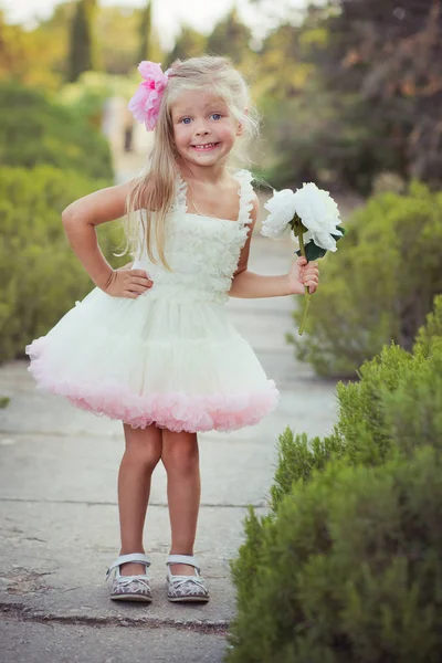 Increíbles ojos azules profundos bebé niña elegante vestido con colorido vestido rosa con pelos rubios brillantes y sandalias blancas posando sentarse para la cámara verano parque central bosque prado con flores y tronco —  Fotos de Stock