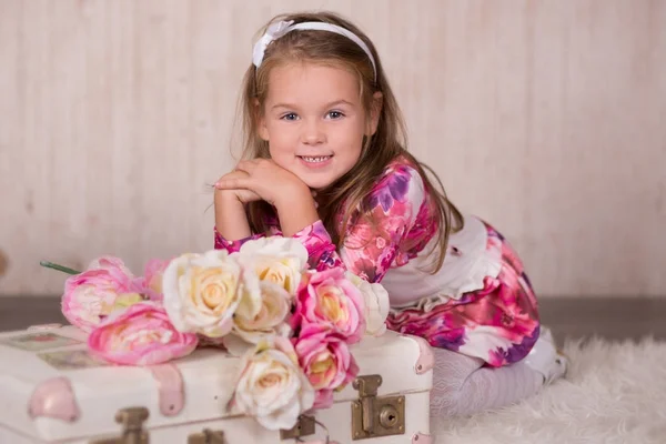 Cute baby girl with brunette hairs pink cheeks and green eyes posing sit for camera in studio wearing airy colourful dress and fancy casuals pumps close to travelling trunk bag with pictures and roses — Stock Photo, Image