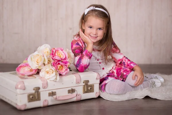 Cute baby girl with brunette hairs pink cheeks and green eyes posing sit for camera in studio wearing airy colourful dress and fancy casuals pumps close to travelling trunk bag with pictures and roses — Stock Photo, Image