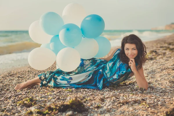 Femme sensuelle incroyable avec des cheveux bruns et des yeux bleus de glace avec des ballons blancs posant assis pour appareil photo sur la plage de sable d'été portant robe colorée modish fantaisie pieds nus — Photo