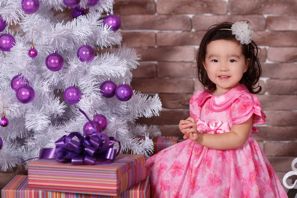 Ásia pin up pouco bonito menina vestindo rosa casual vestido posando perto de novo ano natal branco árvore com violeta roxo bolas brinquedos e coloridos presentes — Fotografia de Stock