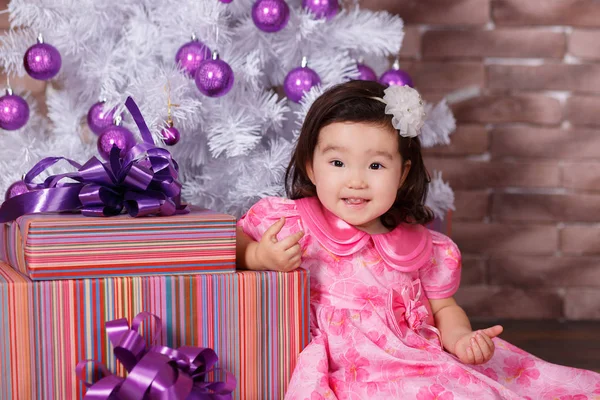 Ásia pin up pouco bonito menina vestindo rosa casual vestido posando perto de novo ano natal branco árvore com violeta roxo bolas brinquedos e coloridos presentes — Fotografia de Stock