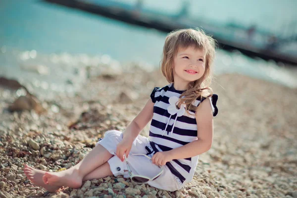 Stijlvol geklede blonde kind meisje geniet van poseren zomervakantie op openbare stad strand dragen casual lichte kleding gelukkig lachend op de camera alleen — Stockfoto