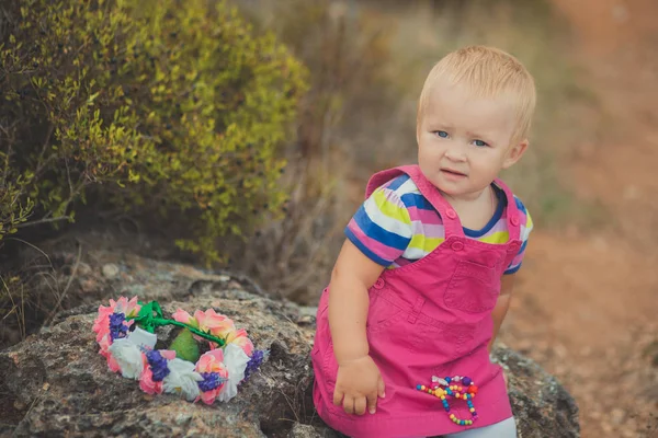 Gelukkig schattige babymeisje met blonde haren dragen van roze jeans jurk poseren glimlachend in het central park tussen groene boom en steen — Stockfoto