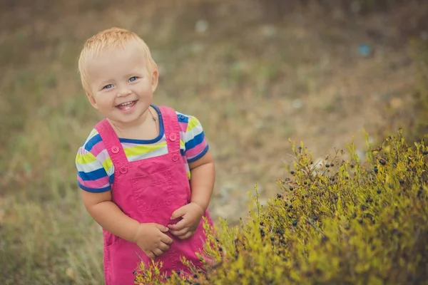 Gelukkig schattige babymeisje met blonde haren dragen van roze jeans jurk poseren glimlachend in het central park tussen groene boom en steen — Stockfoto