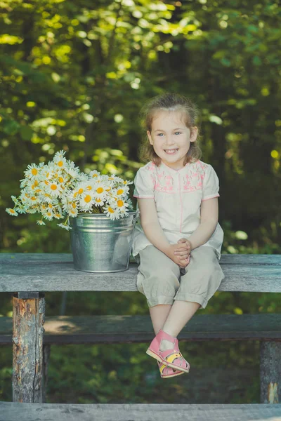 Schattige blonde meisje kind stijlvol gekleed in een wit overhemd en een korte broek zittend op houten bankje poseren emmer vol met daisy kamille mayweed. Adoreable scène — Stockfoto