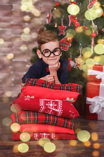 Bonito menino bonito celebrando o Natal de Ano Novo sozinho perto da árvore de xmas no travesseiro vermelho posando na decoração do estúdio vestindo jeans e camisa azul com gravata jazzbow — Fotografia de Stock