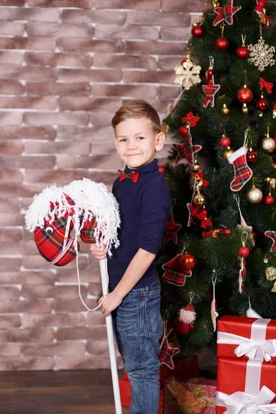 Bonito menino bonito celebrando o Natal de Ano Novo sozinho perto da árvore de xmas no travesseiro vermelho posando na decoração do estúdio vestindo jeans e camisa azul com gravata jazzbow — Fotografia de Stock