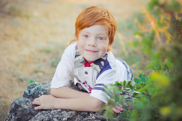 Schöner kleiner Junge mit roten Haaren und blauen Augen, der im Sommer auf der Waldwiese vor der Kamera steht und das warme Klima draußen in der Natur genießt — Stockfoto