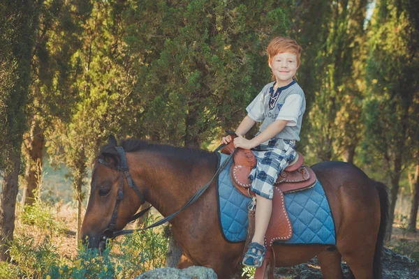 Menino bonito com cabelo vermelho e olhos azuis brincando com seu amigo cavalo pônei na floresta.Enorme amor entre criança shild e animal de estimação fazenda — Fotografia de Stock