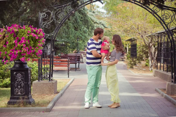 年轻的父亲母亲和婴儿的女儿的家庭在夏季城市的街道公园散步和拥抱, 靠近花束和木凳 — 图库照片