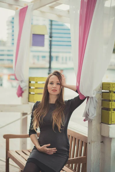 Beautiful young pregnant lady woman with brunette hair lonely dreaming sitting on wooden beanch in waterfront autumn city looking to camera and tender holding her tummy — Stock Photo, Image