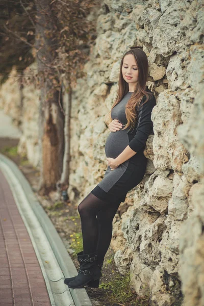 Mulher grávida bonita jovem com cabelo morena solitário sonhando olhos fechados dependem de cerca de pedra parede outono cidade concurso segurando por mãos sua barriga bebê por nascer — Fotografia de Stock