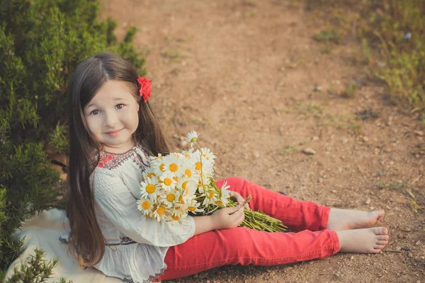 Girl with brunette hair and brown eyes stylish dressed wearing rustic village clothes white shirt and red pants on belt posing with flowers in forest meadow — Stock Photo, Image