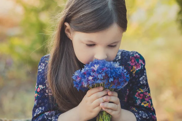 Aranyos kislány és barna haj barna szem portré, mély kék lila vad virágok mezítláb, elegáns színes virág ruhát visel, és szeretnének kamera csillogó arca — Stock Fotó