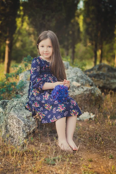 Aranyos kislány és barna haj barna szem portré, mély kék lila vad virágok mezítláb, elegáns színes virág ruhát visel, és szeretnének kamera csillogó arca — Stock Fotó